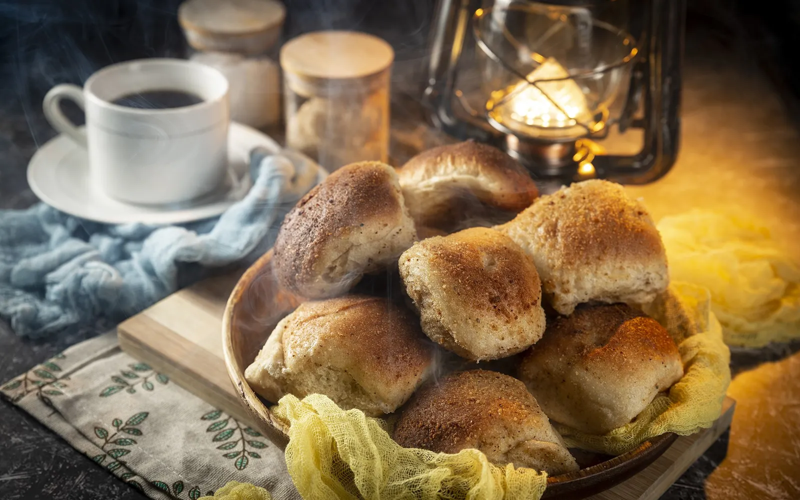 A bowl of fresh pandesal beside a cup of coffee and a lit lantern